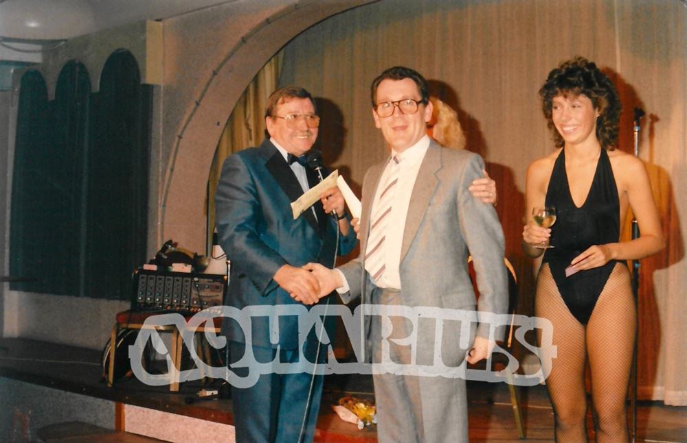 Compere Tufty Gordon on stage with customer, in front of two female staff