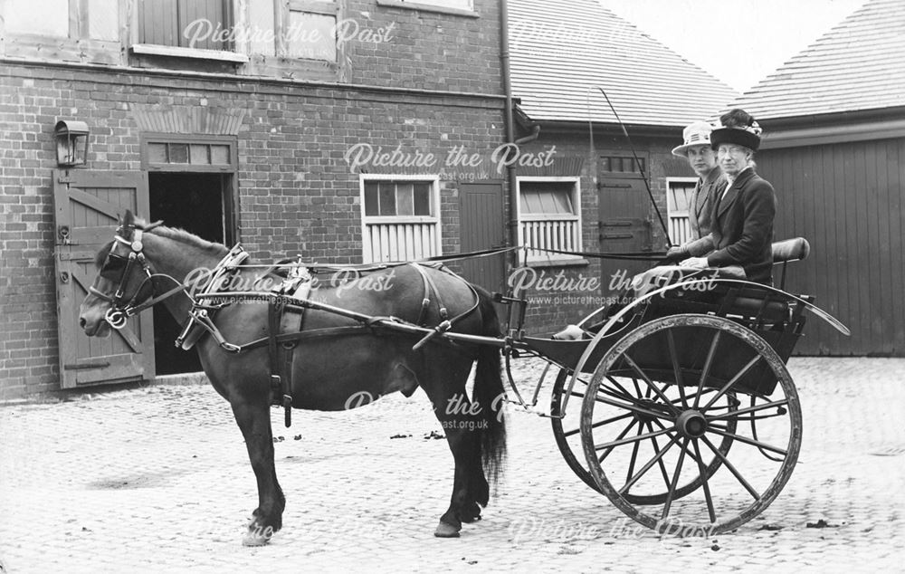 Two ladies in a pony and trap, outside stables